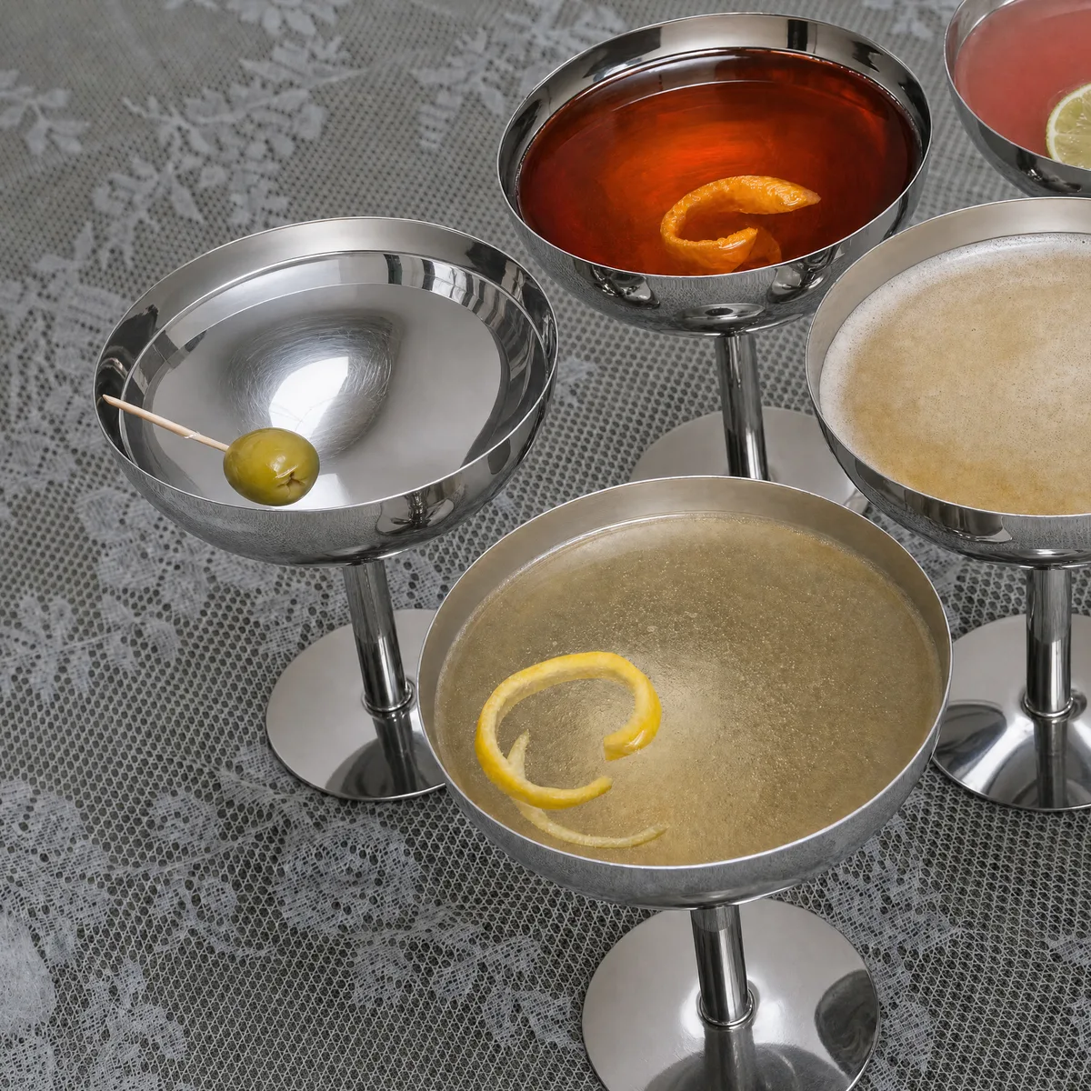 Vijf Vera coupes van rvs met cocktails, een olijf en citrusschil op een grijs kanten tafelkleed