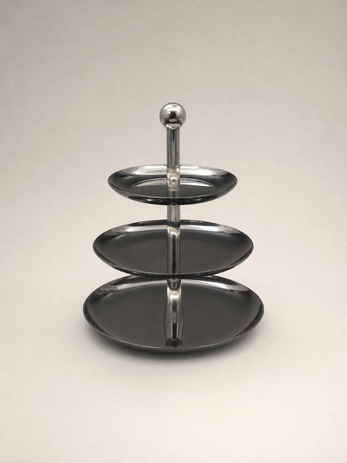 Three-tier polished stainless steel cake stand on a light warm-grey background
